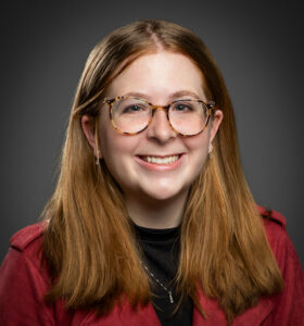Head-and-shoulders portrait of Kira Arkins smiling at the camera. She has long red hair parted down the middle and tucked behind her ears, and she is wearing glasses. She is dressed in a black T-shirt layered under a red top. The background is a muted gray, keeping the focus on her face.