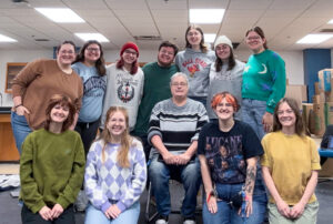 Twelve people pose for a group photo in what appears to be a classroom. Seven people stand in the back row, while five are in the front. Four people in the front row are kneeling, and the person in the center is seated in a chair. Kira Arkins is second from the left in the front row, wearing a purple-and-white argyle-patterned top. Everyone is dressed casually and smiling at the camera. A large stack of cardboard boxes is visible on the right side of the room, while tables and shelving can be seen on the left.