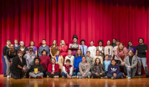 Thirty-four people pose for a group photo on a stage with a shiny wood floor and a red curtain backdrop. Most of the group appears to be middle school students, with a few college-aged individuals standing or kneeling along the edges. At the center of the back row, one person wears a black top hat and holds a large prop chocolate bar labeled “Wonka Bar.” Kira Arkins is kneeling on one knee on the right side of the photo, wearing jeans, a black T-shirt, and a gray button-up shirt. The group is smiling as they celebrate what appears to be a production of Willy Wonka Jr.