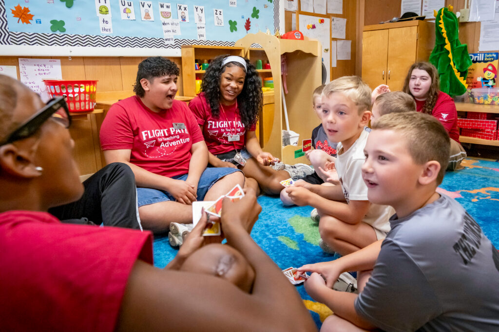 Four Flight Path to Teaching Adventure participants sit on the floor of a classroom, playing an animated game of Uno with several elementary-age children. The participants wear red Flight Path to Teaching T-shirts featuring the program name and a paper airplane design, along with name tags. In the background, a bulletin board displaying the alphabet and shelves filled with toys, games, and classroom supplies suggest a learning environment for young children.