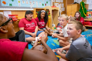 Four Flight Path to Teaching Adventure participants sit on the floor of a classroom, playing an animated game of Uno with several elementary-age children. The participants wear red Flight Path to Teaching T-shirts featuring the program name and a paper airplane design, along with name tags. In the background, a bulletin board displaying the alphabet and shelves filled with toys, games, and classroom supplies suggest a learning environment for young children.