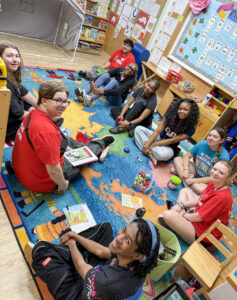 Nine Flight Path to Teaching Adventure participants sit in a circle on a classroom carpet, smiling at the camera. Children's books, containers of toy cars, and toy bugs are placed on the floor in front of them, suggesting they are preparing for an activity. The classroom is filled with brightly colored learning materials and decorations.