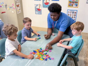 A Flight Path to Teaching Adventure participant sits at a table with three elementary-age children, engaging in an activity with brightly colored toy bugs spread across the tabletop. The participant reaches toward one of the bugs while each child has several in front of them. Children's artwork hangs on the classroom wall behind them, alongside a careers poster, butterfly and insect magnets, and Disney princess magnets displayed on metal cabinets.