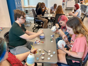 Nine Flight Path to Teaching Adventure participants sit at two tables, painting pottery with colorful glazes. Four participants work at the table in the foreground, while five paint at a table behind them. Small containers of glaze and paintbrushes are spread across the tables, with each participant focused on decorating a pottery piece. In the background, part of another person is visible painting pottery at a separate table. Lockers and worktables line the room, suggesting a classroom or studio space.