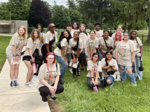 Group photo of the 19 Flight Path to Teaching Adventure participants posing outdoors on a grassy area beside a paved path. The junior and senior high school students wear matching tan T-shirts featuring the Ball State name, the Flight Path to Teaching Adventure logo, and graphics including a compass, schoolhouse, magnifying glass, tent, and camera. Each participant is also wearing a name tag. Trees, a stone wall, and a cross are visible in the background, indicating the photo was taken at Hazelwood Christian Preschool, one of the program's host sites.