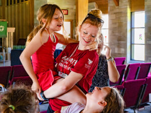 A Flight Path to Teaching Adventure participant holds a smiling elementary-age girl in her arms while another elementary-age girl looks up at them, smiling. The participant wears a red Flight Path to Teaching Adventure T-shirt with a Ball State Teachers College logo on the sleeve, a name tag on a lanyard, and sunglasses resting on top of her head. Her long red hair is pulled into a bun. The children and participant are dressed in warm-weather clothing, and they stand in the aisle of a room with rows of chairs and a podium visible in the background, while other people are present but out of focus.