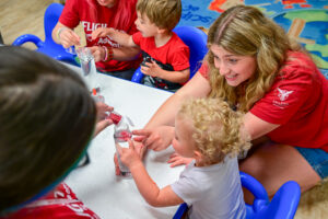 Three Flight Path to Teaching Adventure participants and two preschool-age children work together on a craft activity at a low table. One participant pours red glitter into a bottle of water while another steadies the bottle and talks animatedly with a child, who points toward the bottle. The visible participant wears a red Flight Path to Teaching Adventure T-shirt with a Ball State Teachers College logo on the sleeve and has long blonde hair. The children sit in small blue chairs while the participants kneel or sit on the floor beside them. A brightly colored classroom carpet fills the background.