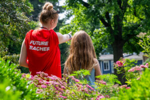 Viewed from behind, a Flight Path to Teaching Adventure participant stands beside an elementary-age child in a garden surrounded by flowers, shrubs, and trees. The participant wears a red Flight Path T-shirt with "FUTURE TEACHER" printed across the back, has her blonde hair in a high bun, sunglasses resting on her head, and points toward something in the distance. The child, with long light brown hair and a blue tank top, looks in the same direction. A blue house is visible beyond the garden.