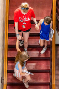 An overhead view shows a Flight Path to Teaching Adventure participant walking down a staircase while holding hands with two elementary-age children. One child walks slightly ahead of the others, and both children carry notebooks as they descend the stairs together.