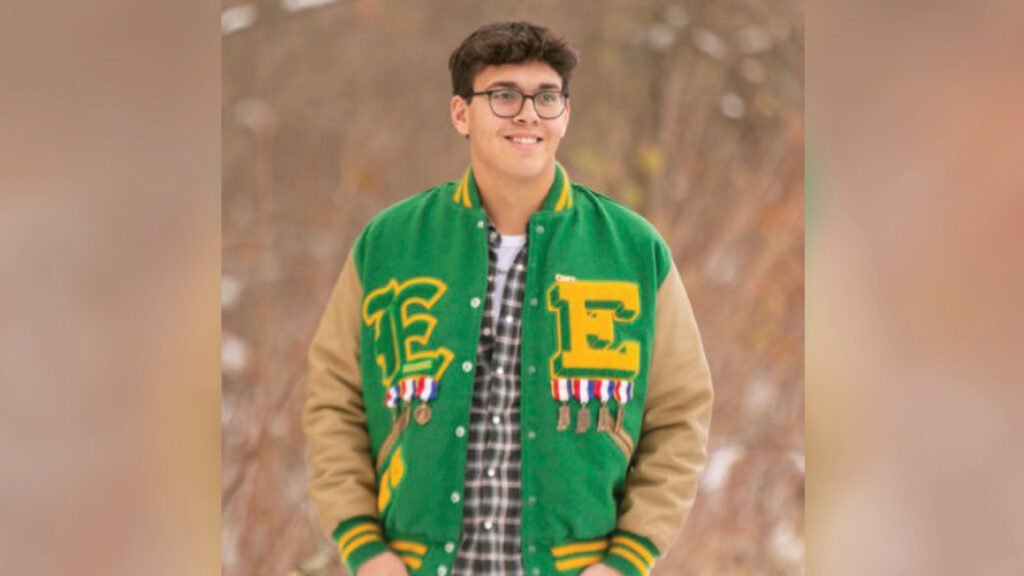 Kaiden Petty stands in a wintery brown field, smiling while looking slightly off to the side. He wears a white T-shirt under a plaid button-up shirt, layered beneath a green and yellow high school letterman jacket. The jacket features large letter "E" patches on both sides, each in a different font. Pinned below the letters are seven or eight Indiana-shaped medals hanging from red, white, and blue striped ribbons. Kaiden has short dark brown hair and wears glasses. The photo has the look and composition of a traditional senior portrait.
