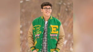 Kaiden Petty stands in a wintery brown field, smiling while looking slightly off to the side. He wears a white T-shirt under a plaid button-up shirt, layered beneath a green and yellow high school letterman jacket. The jacket features large letter "E" patches on both sides, each in a different font. Pinned below the letters are seven or eight Indiana-shaped medals hanging from red, white, and blue striped ribbons. Kaiden has short dark brown hair and wears glasses. The photo has the look and composition of a traditional senior portrait.