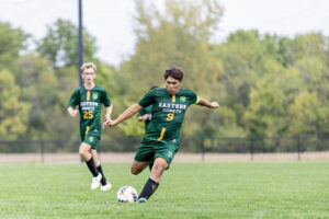 Three Eastern High School soccer players compete on a soccer field. Two players are fully visible, while the arm of a third player appears in the background. They wear dark green uniforms with yellow details, white “Eastern Comets” lettering, yellow numbers, and tall black socks. In the foreground, Kaiden Petty runs toward the ball, preparing to kick it with one foot raised behind him. His arms are extended for balance as he looks down at the ball with a focused expression. Out-of-focus trees are visible beyond a black chain-link fence in the background.