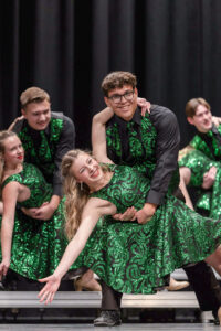 Three pairs of high school students perform on stage during a show choir performance. Each pair features a boy dipping a girl in a choreographed dance pose. In the foreground, Kaiden Petty is in focus, smiling toward the camera as he supports his dance partner in a deep dip. The boys wear black collared dress shirts, black dress pants and shoes, along with vests and ties featuring green glittery swirls on a black background. The girls wear matching dresses made from the same patterned material. Each girl has one arm wrapped around her partner’s neck for support and the other arm extended outward with fingers spread in a jazz hand pose.