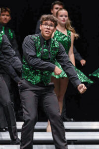 Kaiden Petty performs with his show choir on stage, wearing a black collared dress shirt, black dress pants and shoes, along with a vest and tie featuring green glittery swirls on a black background. He stands in the foreground while several other participants are visible on risers in the background wearing matching outfits. Kaiden holds both hands in fists down to his left side in a choreographed dance move that is mirrored by the performers behind him. His mouth is slightly open, suggesting he is singing or has just finished singing, and he looks toward his right.