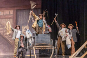 A photograph captures a stage scene from a high school musical theatre performance with a Western theme. Seven performers appear on stage, though five are clearly visible. The performers wear Western-inspired costumes, including pants, shirts, sport coats, and hats. Three performers stand on a wooden wagon, including Kaiden Petty, who is holding a shovel above his head. The other performers hold props including a rope, blanket, garden rake, ax, and rifle, while also carrying blankets. The group appears to be posing at the end of a musical number. The stage backdrop is painted to resemble the staircase of an old saloon or Western building and includes a large “WANTED” poster with a drawing of a person.