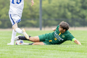 Kaiden Petty plays soccer during a high school game, wearing a dark green Eastern Comets uniform with yellow details, white "Eastern Comets" lettering, yellow numbers, tall black socks, and white cleats. He is stretched out on the ground after making a feet-first slide tackle, with both feet on the soccer ball. The lower half of an opposing player is visible nearby, wearing a white uniform with blue stripes and blue numbers, with one foot drawn back as if preparing to kick the ball before Kaiden's challenge. Out-of-focus trees and a black chain-link fence fill the background.