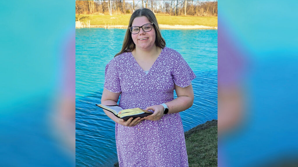 Anna Brewer stands outdoors beside a pond, smiling at the camera while holding an open book with both hands. She wears a floral dress, has long brown hair, and wears glasses. Bare trees line the opposite bank of the pond in the background. Although the trees are bare, the bright sunshine and Anna's floral dress give the scene a springtime feel. The photo appears to be a posed portrait.
