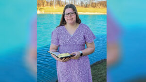 Anna Brewer stands outdoors beside a pond, smiling at the camera while holding an open book with both hands. She wears a floral dress, has long brown hair, and wears glasses. Bare trees line the opposite bank of the pond in the background. Although the trees are bare, the bright sunshine and Anna's floral dress give the scene a springtime feel. The photo appears to be a posed portrait.
