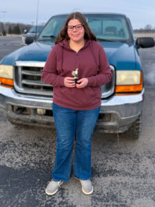 Anna Brewer stands in front of a parked pickup truck, smiling as she holds a small trophy with both hands. She wears jeans, a burgundy hooded sweatshirt, glasses, and has long brown hair. The trophy has a black base topped with a gold-colored figure. Behind her is a large, mostly empty parking lot with one other pickup truck parked nearby.