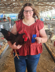 Anna Brewer stands in a 4-H barn at the Jay County Fairgrounds, holding a black rooster in one arm and a blue first-place 4-H ribbon in the other hand. She wears jeans, a red blouse, and glasses, and has long brown hair. Rows of animal cages extend behind her, with a few people standing near other cages in the background.