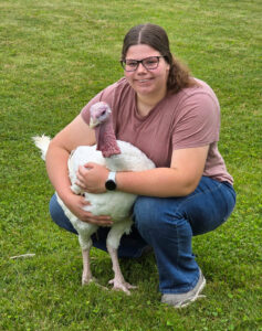 Anna Brewer squats in a grassy area while posing with a large white turkey. She wraps both arms around the turkey in an almost hugging gesture. Anna has long brown hair pulled back in a low ponytail, wears glasses, and is dressed in jeans and a mauve T-shirt. Grass fills both the foreground and background of the photo.