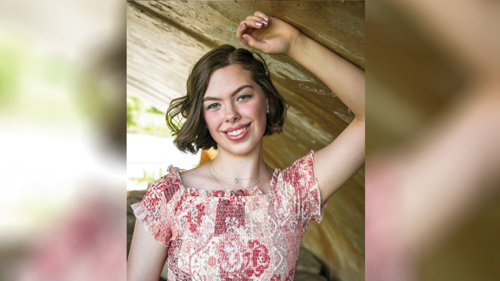 Madelyn Miller poses for a senior portrait-style photo, leaning against her left arm, which is braced on the underside of a concrete tunnel or bridge. Her hand rests above her head as she looks toward the camera. She wears a floral-patterned shirred dress and has brown hair styled in a wavy bob.