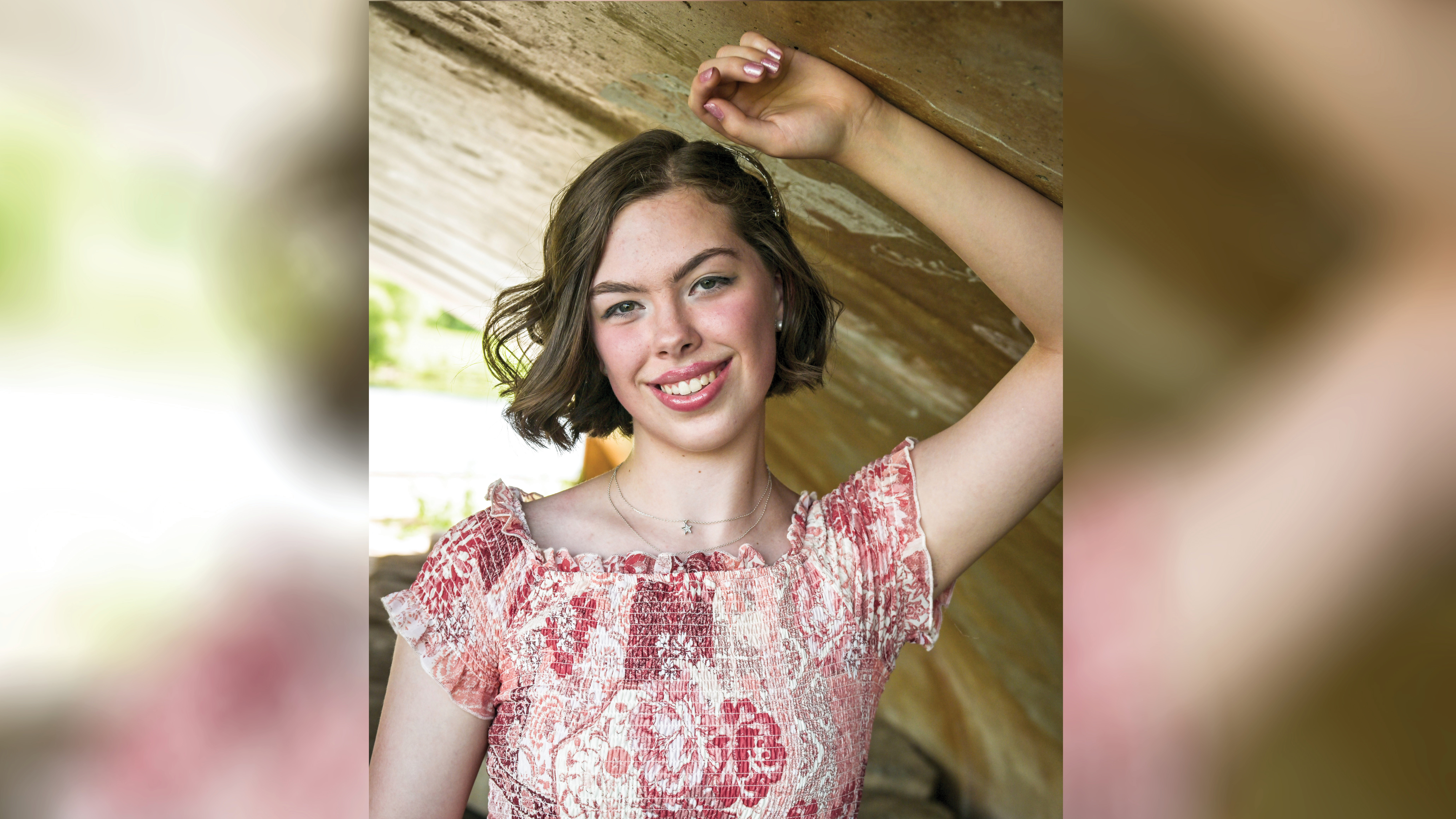 Madelyn Miller poses for a senior portrait-style photo, leaning against her left arm, which is braced on the underside of a concrete tunnel or bridge. Her hand rests above her head as she looks toward the camera. She wears a floral-patterned shirred dress and has brown hair styled in a wavy bob.