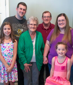 A young Madelyn Miller poses with her family in a multigenerational family photo. Madelyn stands on the left with long brown hair and wears a white sundress decorated with pink, purple, and aqua flowers. Her younger sister Ava stands on the right wearing a pink and salmon striped dress, with her hair pulled back from her face. Several generations of family members stand together, including older and middle-aged relatives. They pose indoors in a warm family portrait setting.