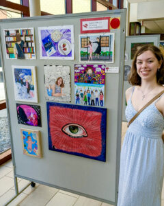 Madelyn Miller poses beside a display board featuring her artwork. She smiles at the camera and wears a pale blue lace dress, a silver necklace with a key charm, small hoop earrings, and a brown crossbody bag. Her shoulder-length brown wavy hair is styled with one side tucked behind her ear. The display board is labeled “Madelyn Miller, 12, AP Drawing, Fishers High School” and features nine colorful drawings and paintings in various sizes and styles, including self-portraits. Other student displays, large windows, and a beige tile floor are visible in the background.