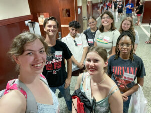 A group of eight young adults take a selfie in a Ball State student center hallway. Madelyn Miller is in the front taking the photo. She has her brown curly hair pulled back and wears a light blue top and a bright pink floral backpack. The group members wear name tags featuring the Ball State Teachers College logo. Many people are visible in the hallway behind them, suggesting a busy campus event such as Orientation or Admitted Student Day.