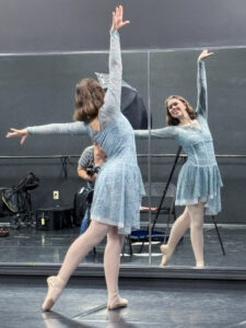 Madelyn Miller poses in a dance studio, with her back to the camera and her reflection visible in a large wall mirror. She smiles as she holds a dance pose with her right arm extended above her head, her left arm stretched out to the side, and her right leg pointed behind her. She wears a light blue long-sleeve dance dress, white tights, and ballet shoes. The studio has black walls, a ballet barre, a folding chair, equipment cases, and a cart visible in the background. The mirror reflection also shows photography equipment and part of the photographer.