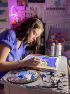 Madelyn Miller paints a canvas on a desk in her bedroom. She holds a thin paintbrush as she adds details to the artwork, with a focused expression. She has brown wavy hair cut in a bob that falls nearly to her shoulders and wears a purple T-shirt with the sleeves rolled up. The desk is covered with a taped-down trash bag and holds a well-used paint palette, tubes of paint, and bottles of black and white paint. In the softly blurred background, her lavender-colored bedroom is visible, including a purple and white bed, a bookshelf with a pink LED light sculpture, and white stylized letters spelling her name on the wall.