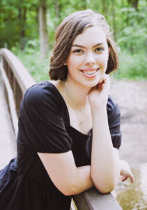Madelyn Miller poses for a traditional senior portrait on a bridge, resting her head on her left hand with her elbow propped on the railing. Her right arm crosses behind her left arm and rests flat along the top of the bridge railing. The bridge appears to cross a small creek on a wooded path, with blurred trees visible in the background. She wears a short-sleeved black dress, two necklaces, and small earrings. Her brown bobbed hair is straightened and parted to the side. The photo has soft, bright lighting that suggests an overcast day.