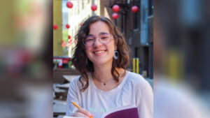 Megan Slack smiles at the camera while holding an open journal with a brightly colored cover that reads “you are loved” in lowercase cursive. She holds a pencil as if writing or drawing. Megan wears a light-colored “be kind” T-shirt, a butterfly pendant necklace, large flat hoop earrings, and round glasses. Her brown wavy layered hair falls below her shoulders. She sits at a picnic table in a courtyard between downtown buildings, with red paper lanterns hanging overhead in the background.