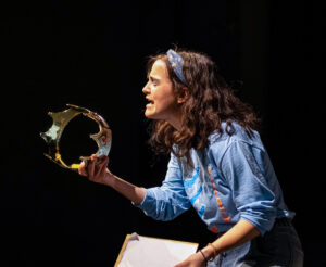 Megan Slack performs on stage under bright overhead lighting against a black backdrop. She holds a gold-colored crown with a jeweled top in one hand and several sheets of paper in the other. Her dark brown wavy hair is pulled back with a blue bandana patterned in yellow. With her eyes closed and mouth open, she appears to be delivering an emotional monologue or singing. She wears a light blue three-quarter-sleeve T-shirt with a blue and orange graphic design.
