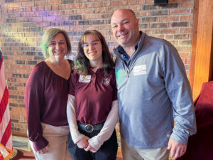Megan Slack poses with two adults, one standing on each side of her, at what appears to be the 2026 Concordia Scholars Breakfast. All three wear name tags, and the adult on one side has a name tag reading "Vince Slack." Megan wears a burgundy Concordia Lutheran High School polo embroidered with the school logo over a white long-sleeve shirt. They smile together in front of a red-and-black brick wall, with part of a United States flag visible on a stand behind them.