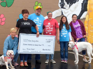 Six people pose in front of a colorful mural with two dogs. Three wear SPCA shirts or sweatshirts, while the other three wear matching shirts that read “Adopt A Fur-Ever Friend” with three paw prints. One person holds a large ceremonial check made out to the Allen County SPCA for $1,173.00. The check is dated September 8, 2019, with “Fur-ever Friends” in the memo line and “Megan’s Mural Project” on the signature line.