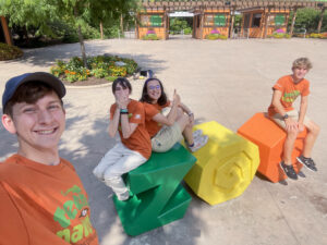 Four young adults wearing matching orange "Teens for Nature" T-shirts and khaki bottoms pose for a selfie at the Fort Wayne Zoo's iconic ZOO letter sculpture. Megan Slack sits on the letter Z with one arm wrapped around her legs and gives a thumbs-up with the other hand. Two others sit on the sculpture, including one laughing with their hands covering their face, while a fourth person stands to the side taking the selfie. The zoo entrance is visible in the background on a sunny day.