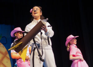 Megan Slack performs on stage wearing a white athletic sweatsuit with black stripe details. Her hair is partially pulled up with a pink flower clipped at the front, and black makeup on her nose suggests she is portraying a dog. She holds a large cardboard rocket with both hands, its nose made from an empty two-liter bottle, while appearing to speak or sing. Two performers in pink cowboy hats and matching pink outfits stand behind her, with a large pineapple prop and a black stage curtain in the background.