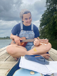 Megan Slack sits cross-legged on a narrow pier overlooking a lake, smiling as she plays a small guitar resting in her lap. She wears denim overall shorts over a navy blue T-shirt, a gray bandana holding back her hair, and round wire-frame glasses. In front of her, a binder of handwritten notes rests on a guitar case, with one blue flip-flop holding the pages open. Her shoes are off, and a yellow butterfly is painted on one of her shins. Trees and the opposite shore of the lake are visible in the background beneath a cloudy sky.