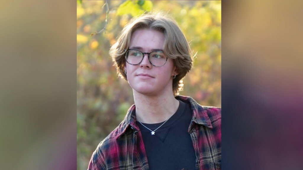 Liam Whitt stands outdoors in soft evening light with a blurred backdrop of trees showing hints of green, yellow, and touches of orange foliage. The photo is framed from the shoulders up, and Liam is looking off to the left rather than at the camera. He has shaggy blond hair that reaches nearly to the bottom of his ears and wears glasses. He is dressed in a black shirt layered with a red, black, and tan plaid overshirt. He also wears a silver necklace with a small star pendant and small hoop earrings in both ears. Liam is holding a book that extends out of the frame, but the visible portion of the cover identifies it as Grimm's Complete Fairy Tales.