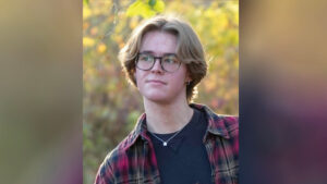 Liam Whitt stands outdoors in soft evening light with a blurred backdrop of trees showing hints of green, yellow, and touches of orange foliage. The photo is framed from the shoulders up, and Liam is looking off to the left rather than at the camera. He has shaggy blond hair that reaches nearly to the bottom of his ears and wears glasses. He is dressed in a black shirt layered with a red, black, and tan plaid overshirt. He also wears a silver necklace with a small star pendant and small hoop earrings in both ears. Liam is holding a book that extends out of the frame, but the visible portion of the cover identifies it as Grimm's Complete Fairy Tales.