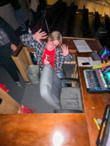 Liam Whitt sits at the sound and lighting booth in a theater, smiling with his hands raised beside his face in a playful "jazz hands" pose while looking off to the side rather than at the camera. The photo appears to have been taken with a camera flash, brightly illuminating Liam and the wooden U-shaped control desk while the theater behind him remains dark. Rows of empty seats are visible in the background, along with dim aisle lighting. On the desk are a soundboard, laptop, open binder, phone resting on papers, and several writing utensils, highlighting the technical work involved in theater productions. Liam wears a red theater-themed T-shirt, though the design is not fully legible, beneath an unbuttoned dark plaid overshirt. Two partially visible people stand in the aisle behind him, cropped by the edge of the frame.