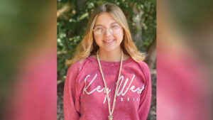 Chloee Ruckman stands outdoors on a sunny day, smiling at the camera. She has long, straight blonde hair and wears round glasses, a red boat-neck sweatshirt with “Key West” in large off-white script and “Florida” in smaller block letters below, and a long beaded necklace with a small potted-plant charm. Out-of-focus trees and foliage are visible in the background.
