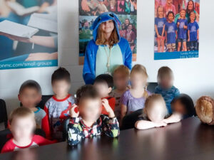 Chloee Ruckman poses with 10 young children in a classroom, all of whose faces are blurred because they are minors. She stands behind the children, smiling and wearing a Stitch costume. The children are arranged in two rows, with some seated at a table and others standing behind them. Posters featuring photos of groups of children hang on the wall in the background.