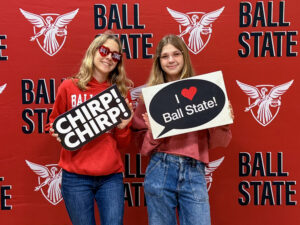 Chloee Ruckman poses with a friend in front of a red Ball State backdrop featuring a repeating pattern of “Ball State” in black text with white shadows and the white Beneficence logo. Chloee holds a sign that reads “I ❤️ Ball State!” while her friend holds a black sign with “CHIRP! CHIRP!” stacked in white text. Both wear red sweatshirts and blue jeans and have long blonde hair. Chloee wears round glasses, while her friend wears red heart-shaped glasses with “Ball” on one lens and “State” on the other.