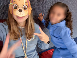 Chloee Ruckman poses in a dog mask and glasses, giving a peace sign with both hands. She wears a muted slate-blue sweatshirt over a white T-shirt and a long beaded necklace with a small potted-plant charm. A student stands beside her, posing with one hand against a carpeted wall. The student has long, curly brown hair and wears a robin’s-egg-blue sweatsuit; her face is blurred because she is a minor. Part of a red block toy is visible in the background, suggesting the photo was taken in a classroom.