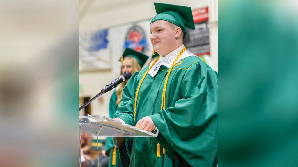 Logan Byrd stands behind a clear podium on stage during his high school graduation ceremony, wearing a green cap and gown with yellow accents, a yellow tassel and cords, and a white graduation stole. Another student stands behind him, out of focus, with signs from the school gymnasium also visible in the background. Logan appears to be giving a commencement speech.