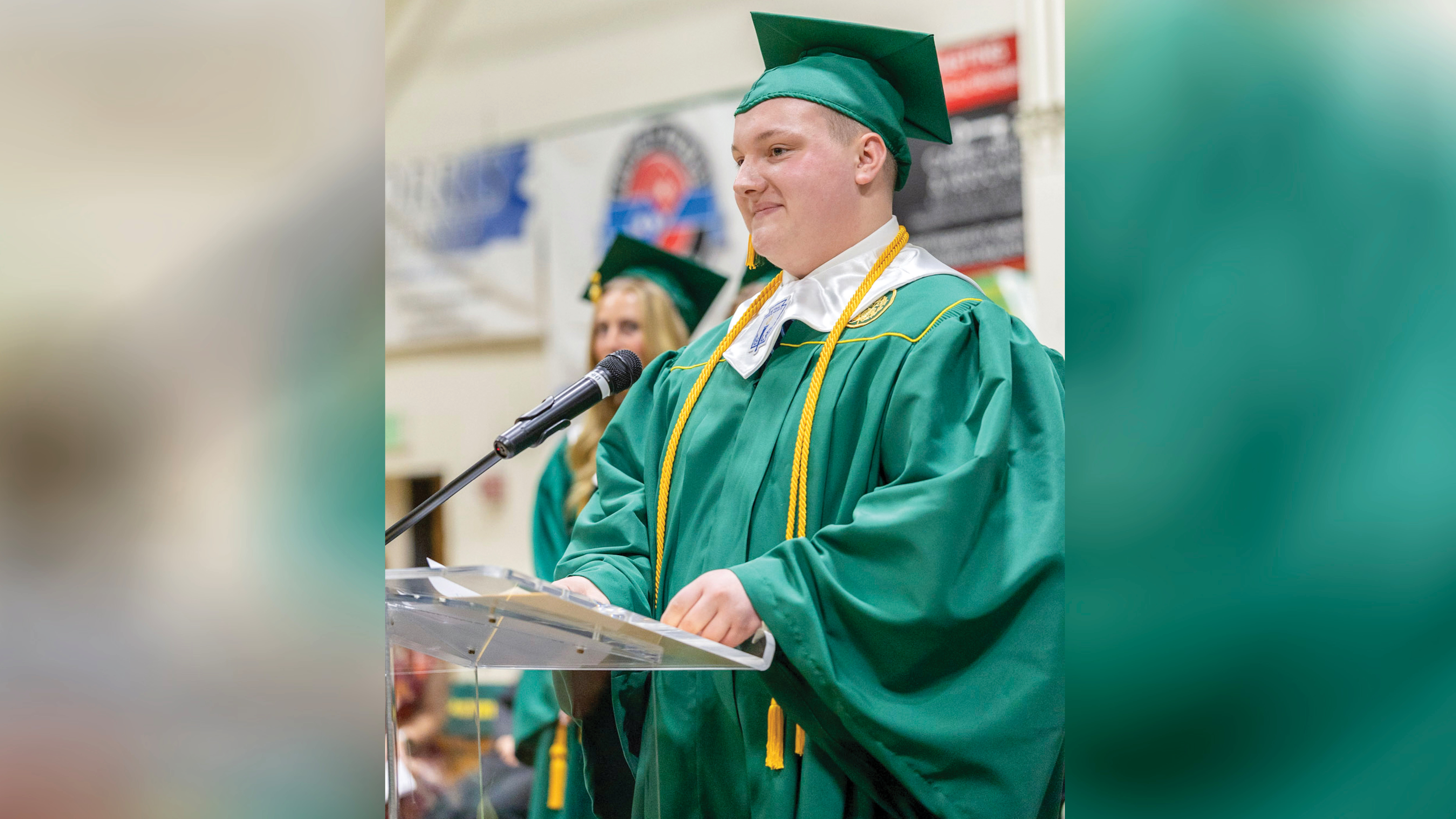 Logan Byrd stands behind a clear podium on stage during his high school graduation ceremony, wearing a green cap and gown with yellow accents, a yellow tassel and cords, and a white graduation stole. Another student stands behind him, out of focus, with signs from the school gymnasium also visible in the background. Logan appears to be giving a commencement speech.