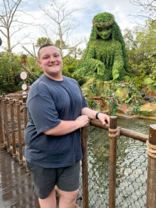 Logan Byrd stands beside a fence at the edge of a pond, smiling at the camera. Behind him is a large topiary resembling Te Fiti from Disney’s Moana. Logan is leaning against the fence and wearing a blue T-shirt and gray pants. The setting appears to be a Disney amusement park.